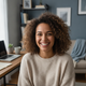 Smiling woman in her late 20s, casually dressed, in a modern home office setting with a laptop.