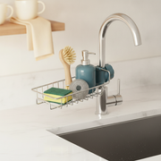 Close-up of a slate blue sink caddy organizer on a bright white kitchen counter, holding a sponge, dish brush, scrubber, and 